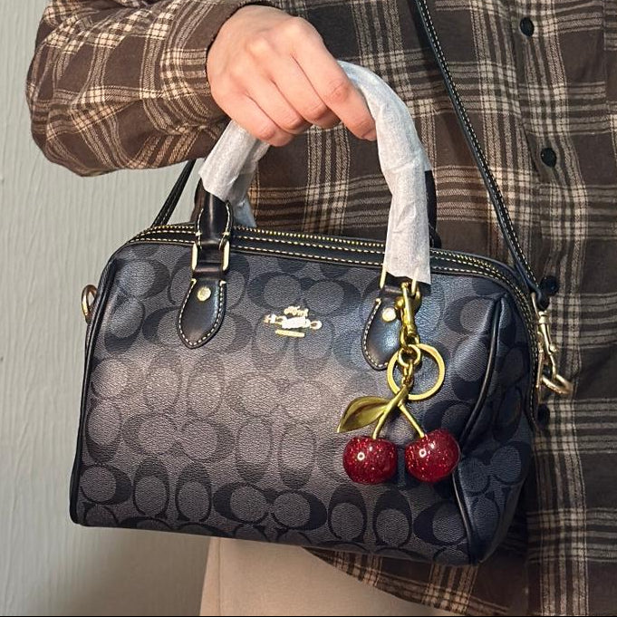 Person holding a Coach handbag with cherry charm against a neutral background